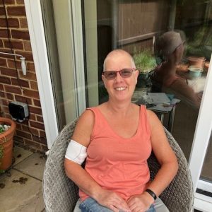 Jo sits on a chair, smiling, with a shaved head.